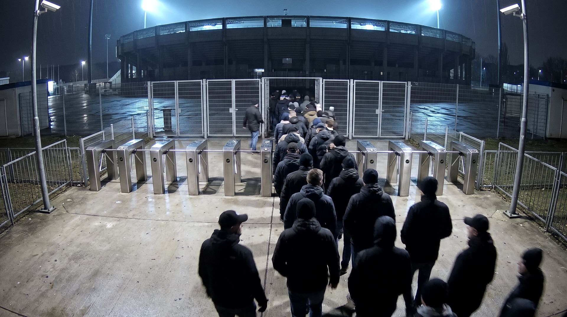Elevated camera view over stadium turnstiles as supporters enter at night